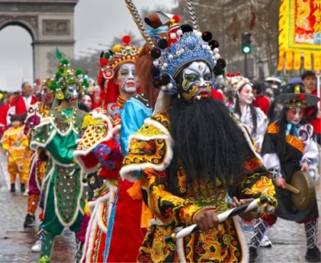Performers in elaborate traditional Chinese opera costumes and painted masks parade down a city street during a festival, with one figure in front wearing a blue headdress, long black beard, and embroidered black-and-gold robe while holding drumsticks.