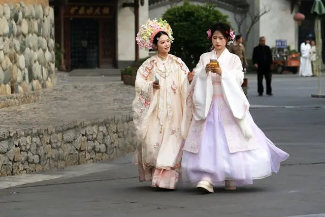 Two women walking outdoors in traditional Chinese Hanfu clothing — one in an embroidered cream robe with an elaborate floral headdress, the other in a white jacket with a flowing lavender skirt — in front of a historic stone-wall building.