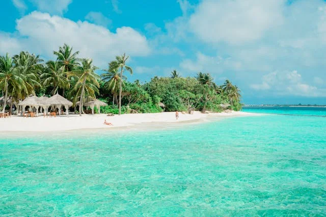 Crystal-clear turquoise water along a tropical white-sand beach lined with palm trees and small thatched huts under a bright blue sky.