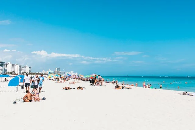 People relaxing and sunbathing on a crowded sandy beach under a clear blue sky near the ocean.