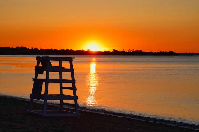 Wooden lifeguard chair silhouetted on a quiet beach at sunset, with orange skies reflecting across calm coastal water.