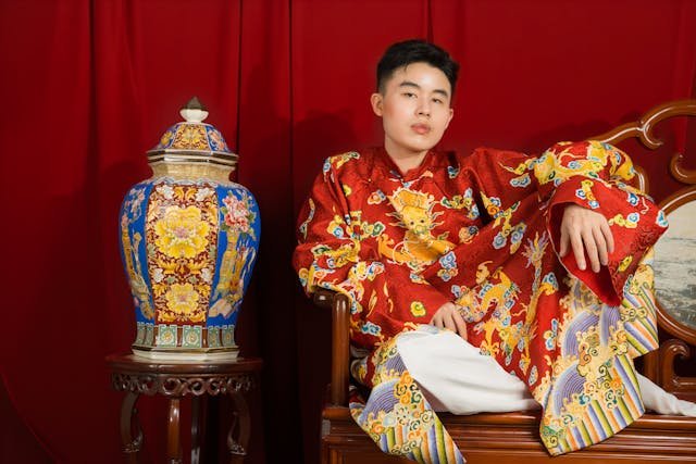 A young person wearing a traditional red embroidered robe sits on a wooden chair beside a decorative ceramic vase against a red curtain backdrop.