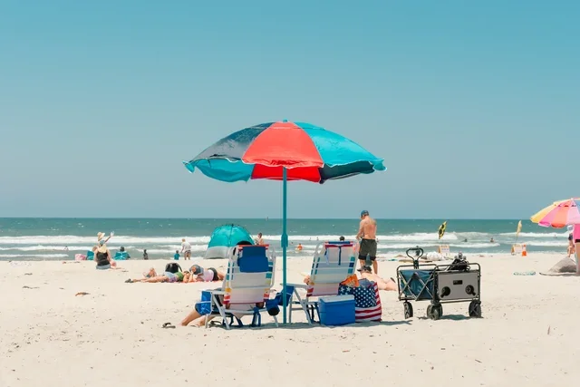 Sunny beach scene with colorful umbrellas, beach chairs, and people relaxing by the ocean on a bright summer day.