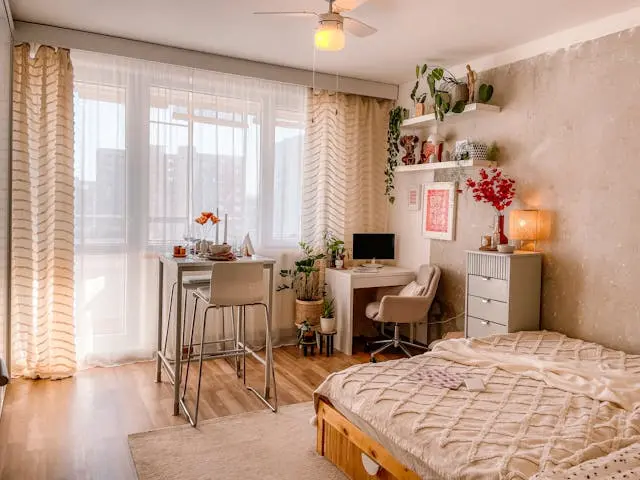Cozy small bedroom with neutral decor, sheer curtains, compact workspace, floating shelves, indoor plants, and a low wooden bed in a bright modern apartment.