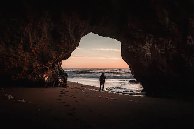 Dark sea cave opening onto a beach at sunset, with a lone person standing near the shoreline and waves rolling in from the ocean.