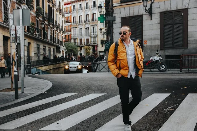 Man wearing a yellow puffer jacket and sunglasses walking across a city crosswalk surrounded by urban buildings and street traffic.