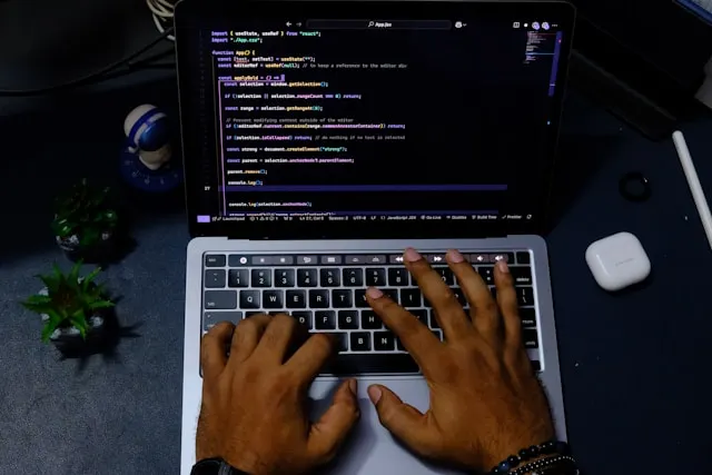 Person typing code on a MacBook laptop with a dark-themed code editor open on screen