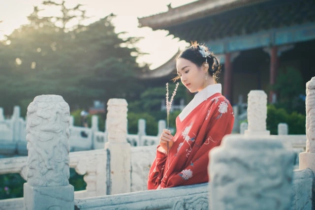 Young woman wearing a red traditional Chinese hanfu robe with white floral embroidery, standing at a stone balustrade in front of a historic Chinese pavilion at golden hour